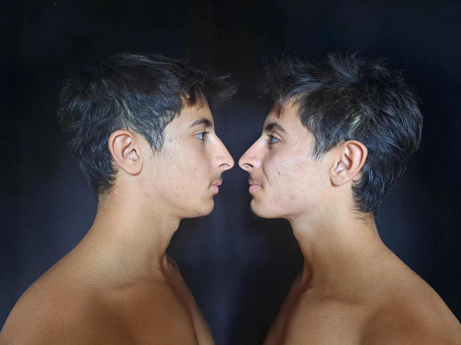 Deux jeunes hommes de profil, face à face, sur un fond sombre.